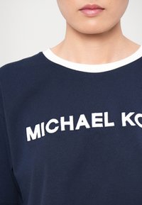 La T-shirt di cotone blu navy presenta un col girocollo bianco e il testo ricamato "MICHAEL KORS" in bianco a contrasto. Tessuto morbido, vestibilità comoda.