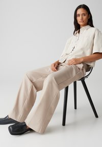 Chemisette beige clair à manches courtes et à deux poches poitrine, assortie à un pantalon large beige clair. Des mocassins noirs sont posés sur une chaise.