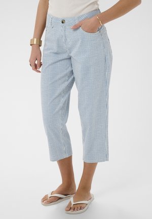 Femme portant un pantalon court rayé vertical bleu et blanc, des tongs blanches, un haut blanc et des bracelets dorés, debout avec une main dans la poche.