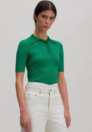 Frau mit dunklen Haaren, die ein grünes, geripptes Poloshirt mit kurzen Ärmeln und hoch taillierte weiße Hose vor neutralem Hintergrund trägt.