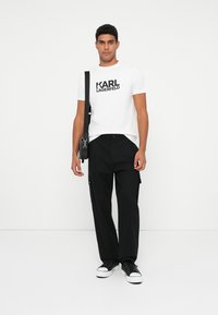 Valge t-särk musta "KARL LAGERFELD" logoga, kombineeritud mustade cargo-pükstega. Modell kannab musti spordijalatseid ja hoiab musta kotti.