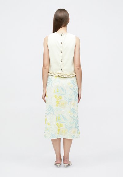 Femme aux cheveux longs et lisses portant un haut crème sans manches avec des boutons dans le dos et une jupe midi fleurie avec un motif bleu clair et jaune.