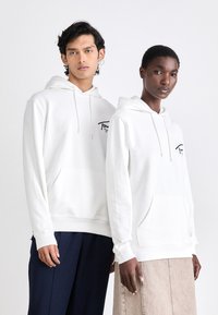 Sudaderas blancas de tejido suave con bolsillos frontales, que presentan un logo impreso cerca del pecho izquierdo. El modelo se combina con pantalones en tonos neutros.