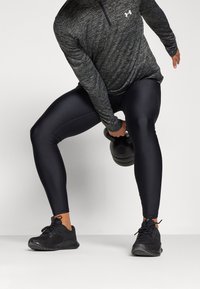 Schwarzes Heather-Langarmshirt mit Viertelreißverschluss, glänzende schwarze Leggings und schwarze Sportschuhe, während sie in Bewegung einen schwarzen Kettlebell hält.