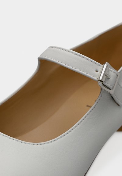 Λευκό δερμάτινο παπούτσι Mary Jane με ασημένιο λουράκι και ταμπά εσωτερική επένδυση, απεικονιζόμενο σε κοντινή γωνία.
