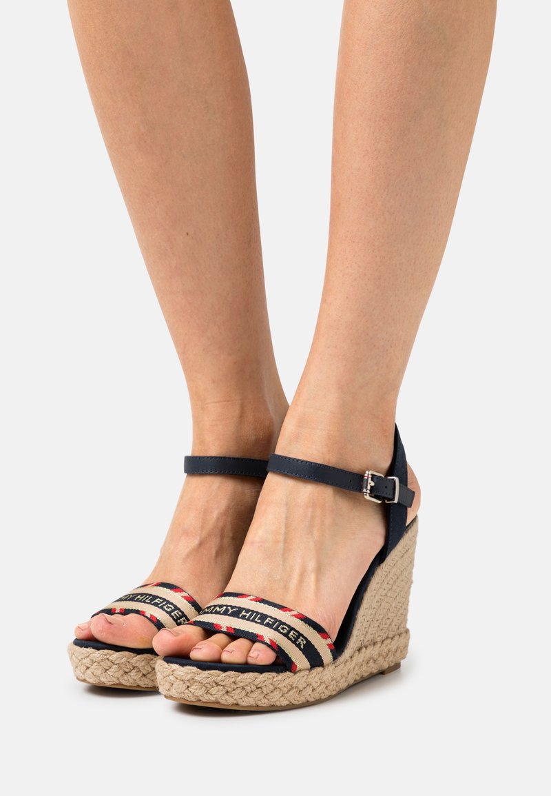 Pieds portant des sandales compensées bleu marine à bride à la cheville, avec semelles en corde tressée et brides avant arborant le logo Tommy Hilfiger et des rayures rouges, blanches et bleues.