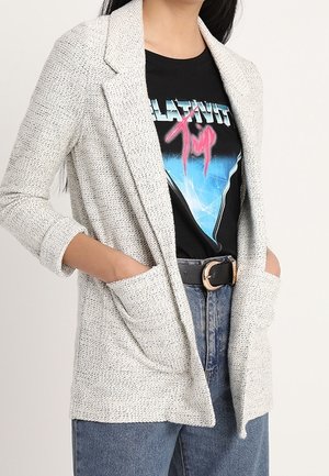 Kobieta ubrana w teksturowaną, jasno szarą marynarkę na graphiczną czarną koszulkę z niebiesko-różowym wzorem, w połączeniu z wysokim stanem niebieskich jeansów i czarnym paskiem.