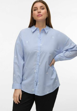 Žena oblečená v světle modré košili na knoflíky s drobnými bílými květinovými vzory a černých kalhotách, pózující s jednou rukou na boku.