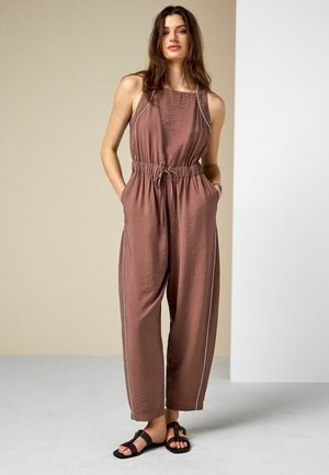 Femeie stă cu mâinile în buzunare, purtând un combinezon maro fără mâneci cu dungi laterale și sandale negre plate, fundal neutru.