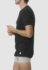 T-shirt nera in cotone a maniche corte, con scollo a V e un logo bianco. Indossata sopra dei pantaloni corti grigi, la modella si trova di profilo su uno sfondo chiaro.