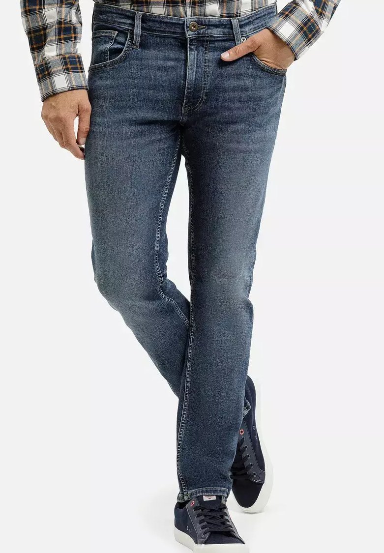 Mężczyzna w dopasowanych niebieskich dżinsach, z jedną ręką w kieszeni, w połączeniu z granatowymi sneakersami i koszulą w kratę.