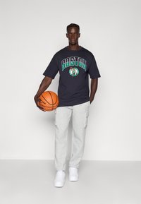 Melna t-kreklā ar "BOSTON" grafiku, pelēkos kargo sporta biksēs, turot oranžu basketbola bumbu, baltajās čībās, stāvot pret pelēku fonu.