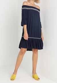 Robe bleu marine à épaules dénudées avec broderies blanches au niveau de l'encolure, ourlet volanté et manches cloches, associée à des mules jaunes à bouts ouverts.