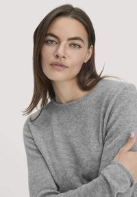 Grauer, langärmeliger Pullover aus weichem, strukturiertem Stoff mit Rundhalsausschnitt und engen Bündchen, getragen von einer Person mit langen Haaren.