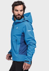 Blaue Softshell-Jacke mit einem kontrastierenden, dunkleren Blauen Paneel, verstellbarer Kapuze, Vorderseite mit Reißverschluss und Logodetails an den Ärmeln.