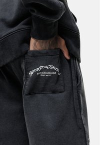 Zwarte sweatshirt met geribbelde manchetten, gecombineerd met donkere broek met een zijzak. De zak toont een wit logo en het jaartal van oprichting.