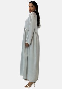 Longue robe gris clair en tissu soyeux avec manches longues et corsage ajusté, présentant une jupe à volants et des fentes latérales, assortie à des talons blancs.