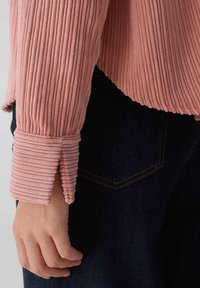 Pink ribbed long-sleeve top with textured fabric and flared cuffs, paired with dark denim. Cuffs feature distinct vertical ridges.
