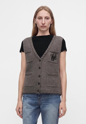 Jeune femme portant un gilet gris tricoté à boutons avec des poches, par-dessus un top noir à manches courtes et un jean bleu, debout devant un fond uni.