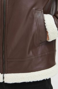 Chaqueta de cuero marrón con un acolchado de piel de oveja blanca en el cuello y el dobladillo, con cierre de cremallera y detalles de bolsillo laterales.