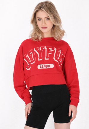 Młoda kobieta o blond włosach nosi czerwoną krótką bluzę z napisem „Joyful League” oraz czarne, wysokie spodenki kolarskie.
