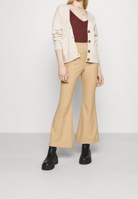 Vrouw in een beige cardigan, maroon tanktop, wijde tan broek en zwarte platformschoenen die tegen een effen witte achtergrond staat.