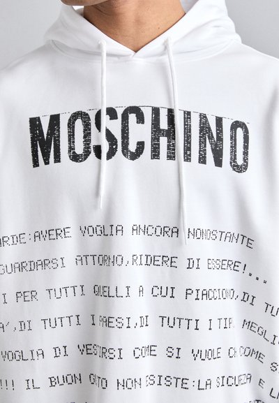 Weiße Kapuzenjacke mit "MOSCHINO" in großen schwarzen Buchstaben am Kragen. Text in unterschiedlichen Größen und Stilen bedeckt die Vorderseite und erzeugt einen markanten grafischen Effekt.