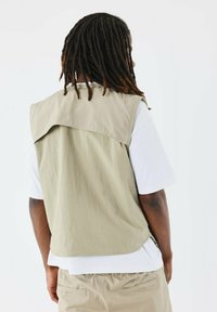 Gilet senza maniche color khaki con un collo alto e un tessuto leggermente texture, sovrapposto a una maglietta bianca, visto da dietro.