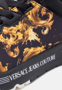 Αθλητικά παπούτσια με μαύρο και χρυσό στροβιλιζόμενο μοτίβο, μαύρα κορδόνια και λευκό λογότυπο "VERSACE JEANS COUTURE" στο πλάι.