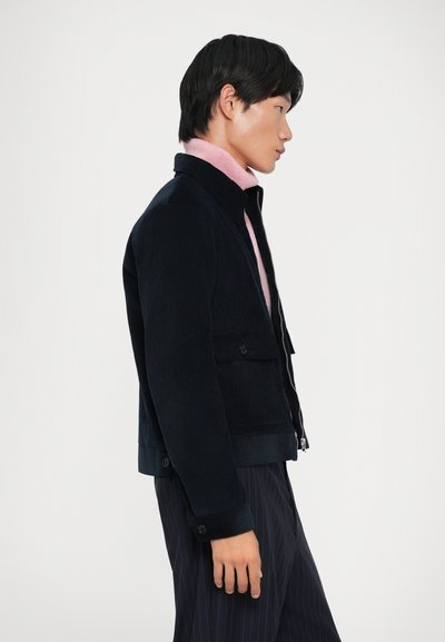 Veste en velours côtelé navy avec fermeture éclair, poches boutonnées et coupe ajustée, superposée à un col roulé rose clair.