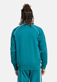 Tyrkysová sportovní bunda s bílými lemy, dlouhými rukávy, žebrovanými manžetami a vysokým límcem. Hladká textura a přiléhavý střih vhodný pro sporty.
