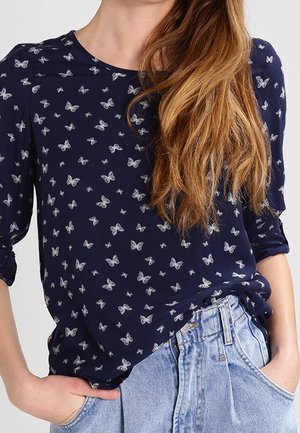 Person trägt ein marineblaues Langarmshirt mit weißen Schmetterlingsmotiven, das in eine hellblaue, hoch taillierte Jeans gesteckt ist, Hände in den Taschen, Haare offen.