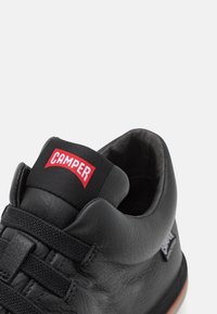 Černá kožená teniska s polstrovaným límcem, pletenými šňůrkami a červeným štítkem "CAMPER" na jazyku. Hladký povrch s bílou podrážkou.
