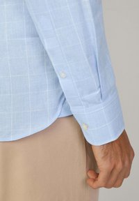 Camisa a cuadros de color azul claro con una textura suave, que presenta un dobladillo curvado, puños abotonados y un patrón de cuadrícula sutil.
