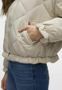 Beige kviltitud bomber-jakk ribatud pitseriga, küljetasku ja köitnud varrukate mansettidega, kombineeritud siniste teksapükstega.