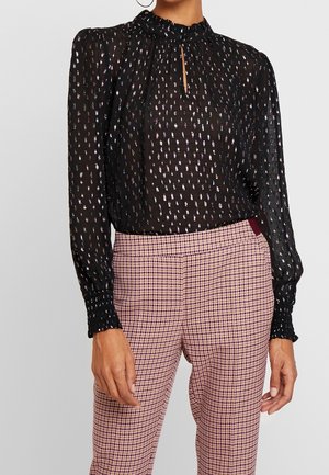 Femme portant un chemisier noir transparent à pois argentés et manches longues, associé à un pantalon taille haute à carreaux roses et bordeaux.