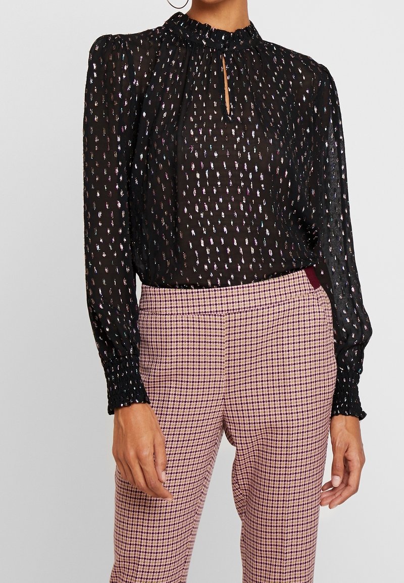 Femme portant un chemisier noir transparent à pois argentés et manches longues, associé à un pantalon taille haute à carreaux roses et bordeaux.