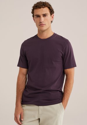 Homme aux cheveux bouclés portant un t-shirt uni violet foncé à manches courtes et un pantalon beige clair, debout devant un fond uni.