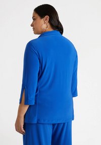 Niebieska bluzka uszyta z gładkiego, lekkiego materiału. Posiada rękawy o długości trzy czwarte z akcentami guzików oraz boczne rozcięcia dla swobody ruchów.