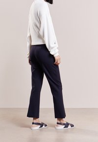 Vit ribbad långärmad tröja, svarta åtsittande byxor och mörka sandaler med bred rem och vit sula, sedd bakifrån.