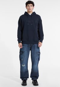 Sudadera de marina con cordones, bolsillo frontal y logotipo; combinada con jeans cargo desgastados con múltiples bolsillos y un ajuste relajado.