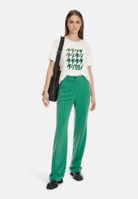 Weißes T-Shirt mit grünem Grafikmuster, kombiniert mit grüner Hose und schwarzen Schuhen. Die Hose hat einen Knopfverschluss und eine gerade Passform.