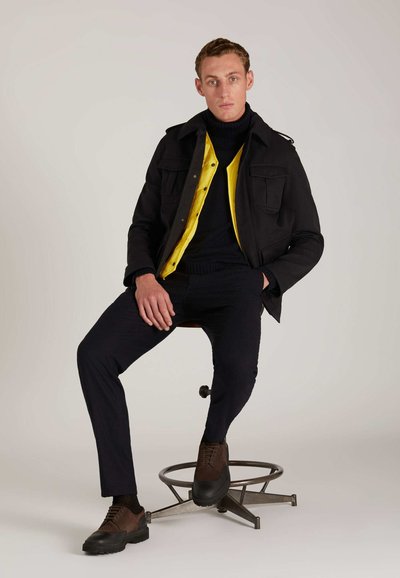 Schwarze Jacke mit zwei Brusttaschen über einem gelben Hemd mit Knöpfen, kombiniert mit einem marineblauen Rollkragenpullover, marineblauen Hosen und braunen Schuhen mit dicken Sohlen.