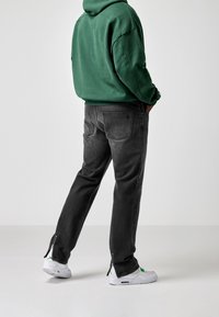 Jeans in denim nero con taglio dritto, tasche posteriori e dettagli con zip sui polsini, abbinati a una felpa con cappuccio verde e sneakers bianche.