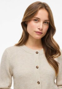 Cárdigan de punto beige con cuello redondo y cierre de botones. La textura es suave, con detalles acanalados y pequeños botones marrones.