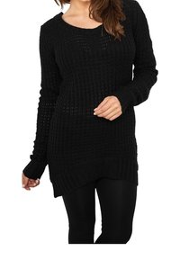 Femme portant un long pull en tricot noir avec un ourlet asymétrique et des leggings noirs, posant une main près de sa taille.