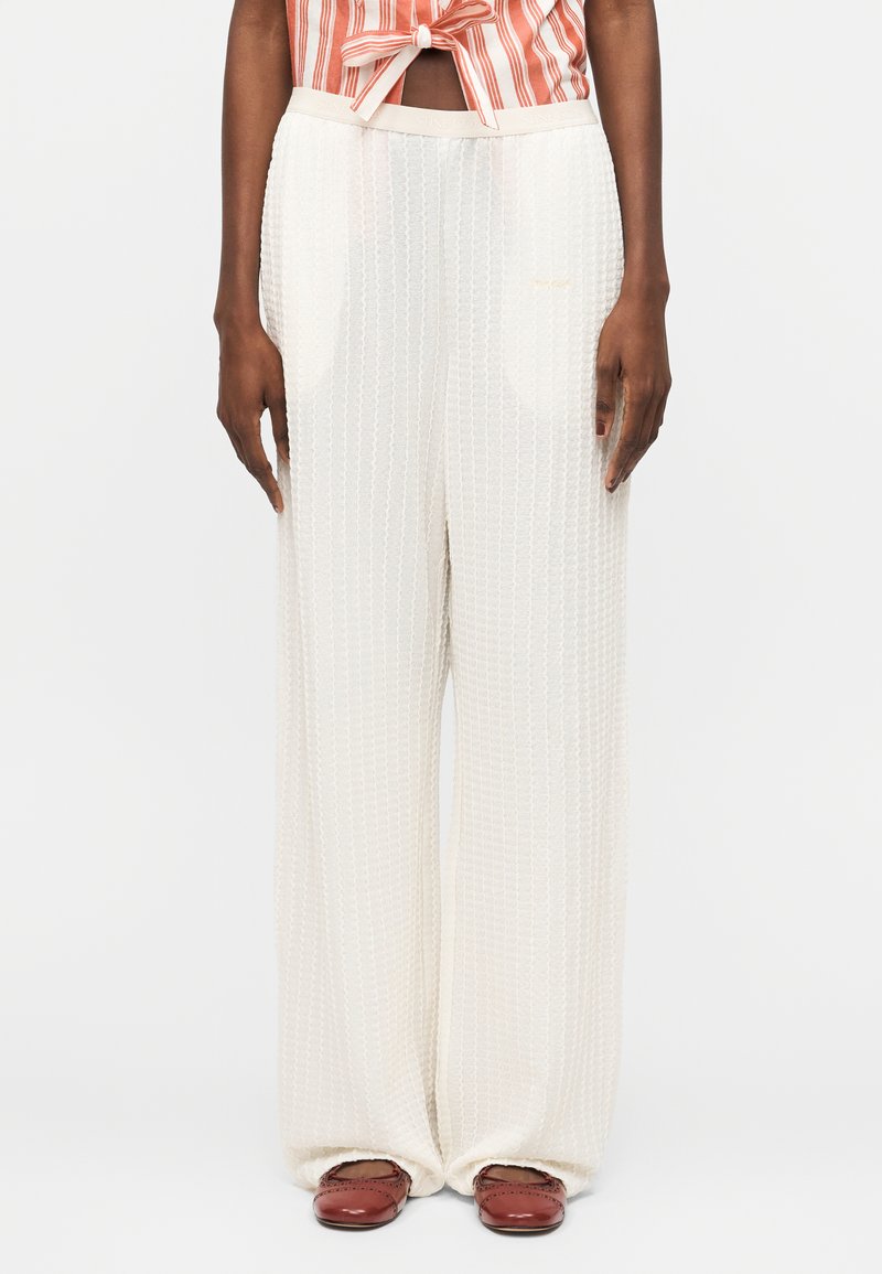 Άτομο που φοράει άνετο λευκό υφασμάτινο παντελόνι, κόκκινο-λευκή ριγέ δεμένη κορυφή και κόκκινα επίπεδα παπούτσια, στέκεται μπροστά σε απλό φόντο.