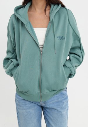 Mujer que lleva una sudadera con capucha turquesa con cremallera y el logo "even & odd" en el pecho, camiseta blanca debajo y vaqueros azul claro con las manos en los bolsillos.