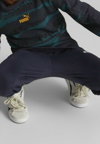 Sportovní outfit skládající se z tmavě modré mikiny se tyrkysovými pruhy a logem Puma, doplněný o tmavě modré tepláky a krémové tenisky.