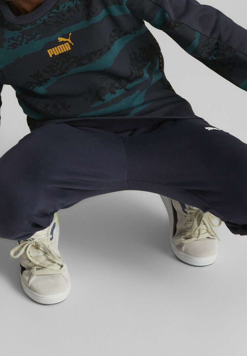 Sportovní outfit skládající se z tmavě modré mikiny se tyrkysovými pruhy a logem Puma, doplněný o tmavě modré tepláky a krémové tenisky.